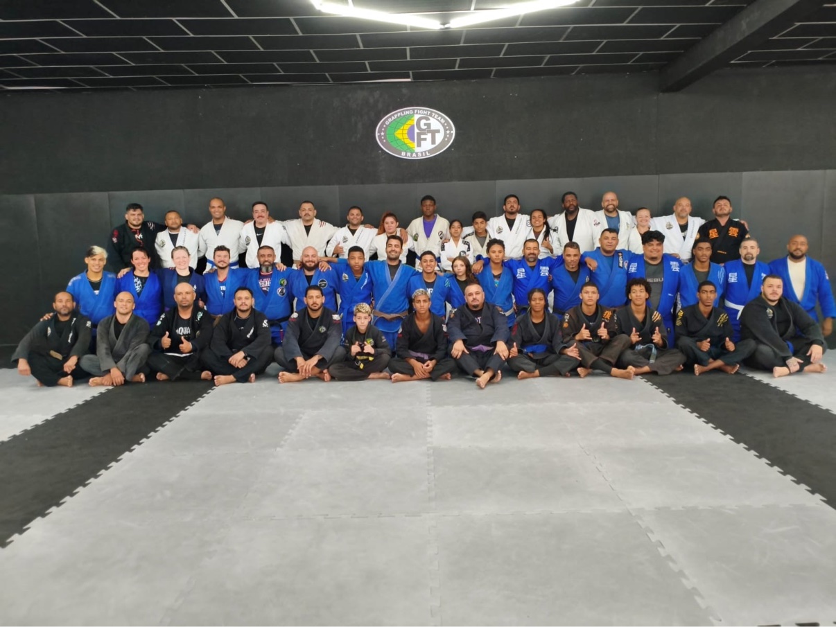 Large group of GFTeam students in blue, white, and black gis posed on the mats under the GFTeam Brasil logo